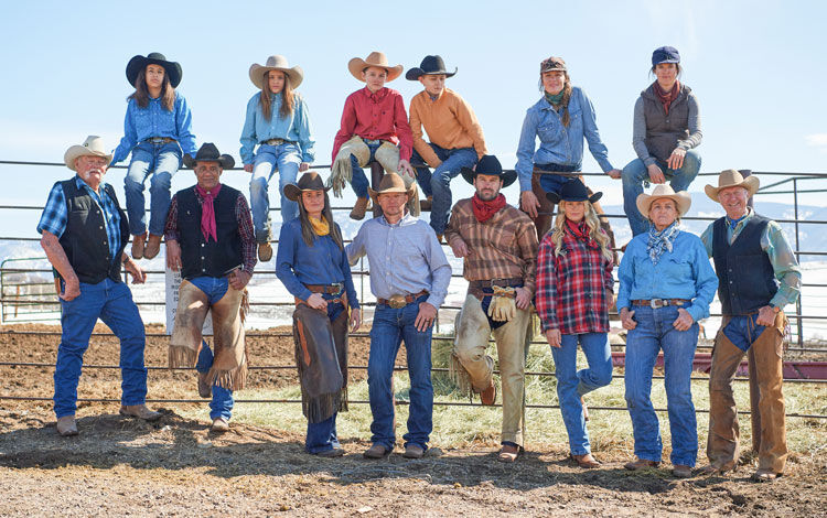 A group of cowboys and cowgirls bundled up in winter clothes and posed for the photo on and around a metal fence.